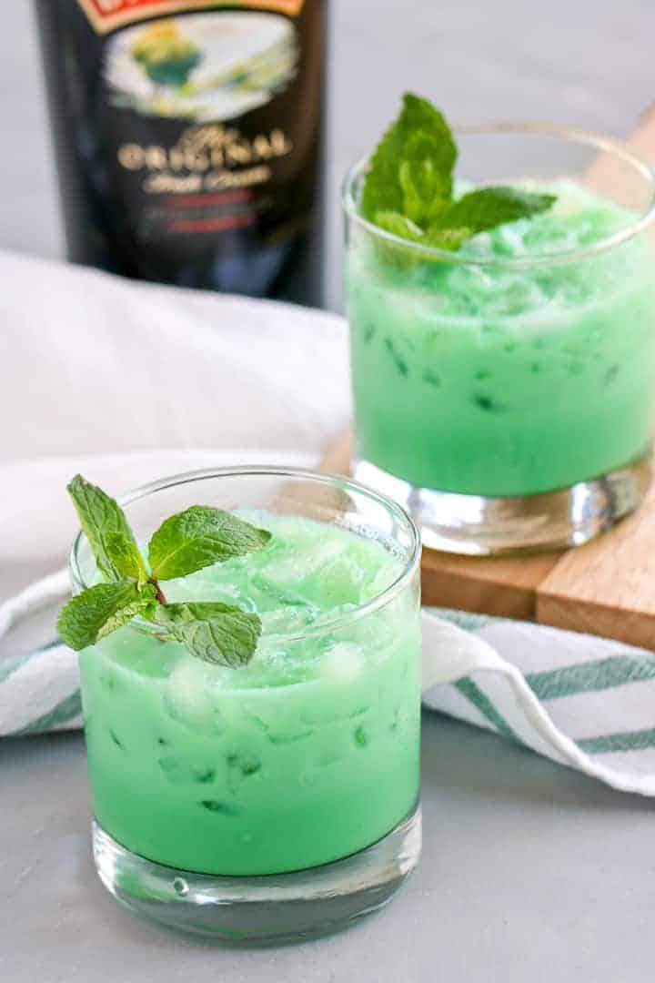 Two glasses of bright green iced drinks, garnished with fresh mint leaves, sit on a table with a striped cloth and a bottle partially visible in the background.