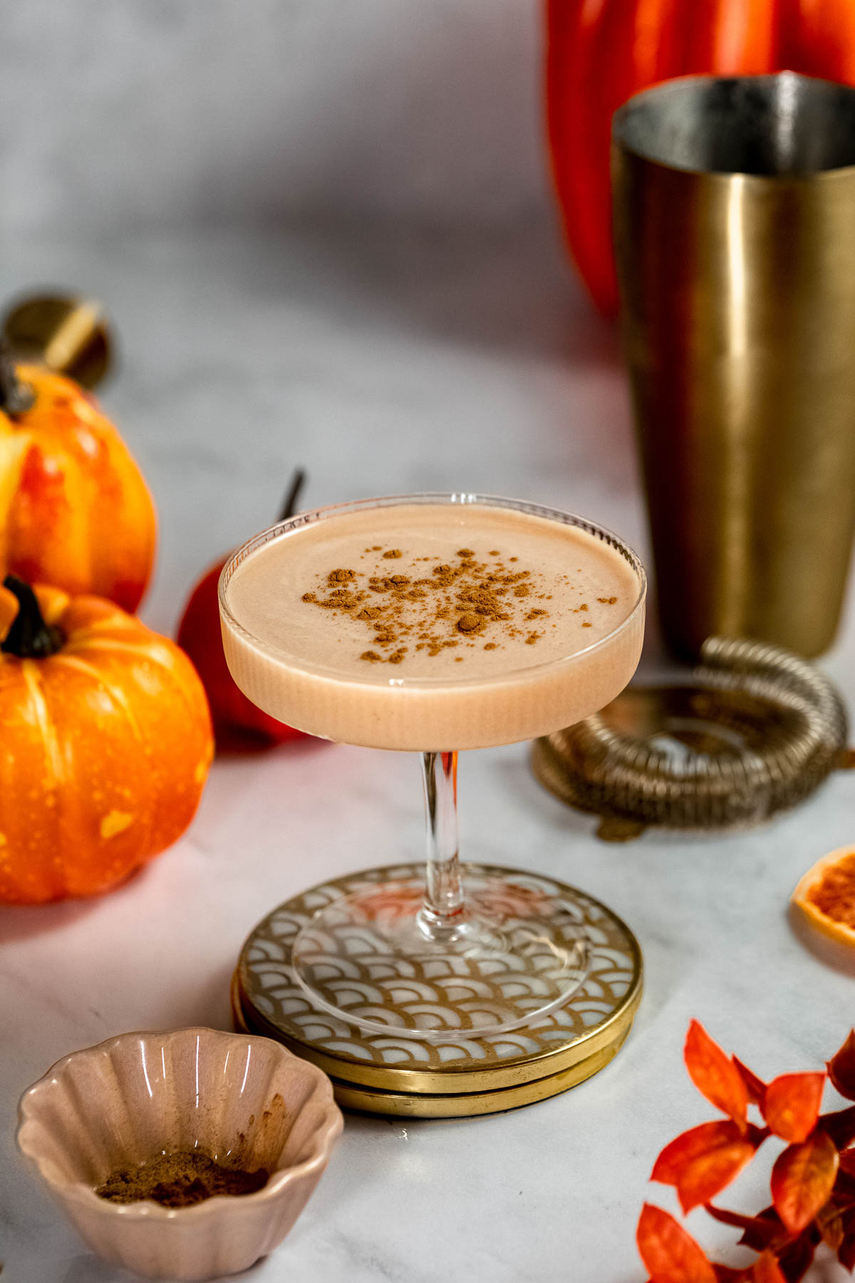 A creamy cocktail garnished with spices in a coupe glass, surrounded by small pumpkins, a cocktail shaker, and autumn decor on a marble surface.