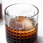 A glass of dark liquor with a large ice cube, set on a white surface.