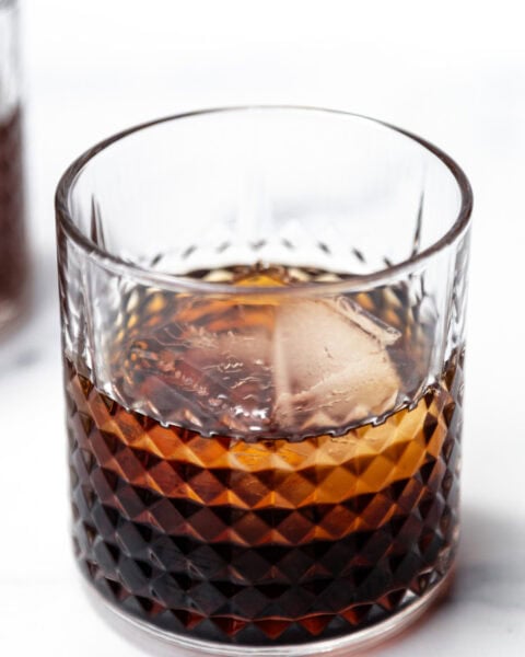 A glass of dark liquor with a large ice cube, set on a white surface.