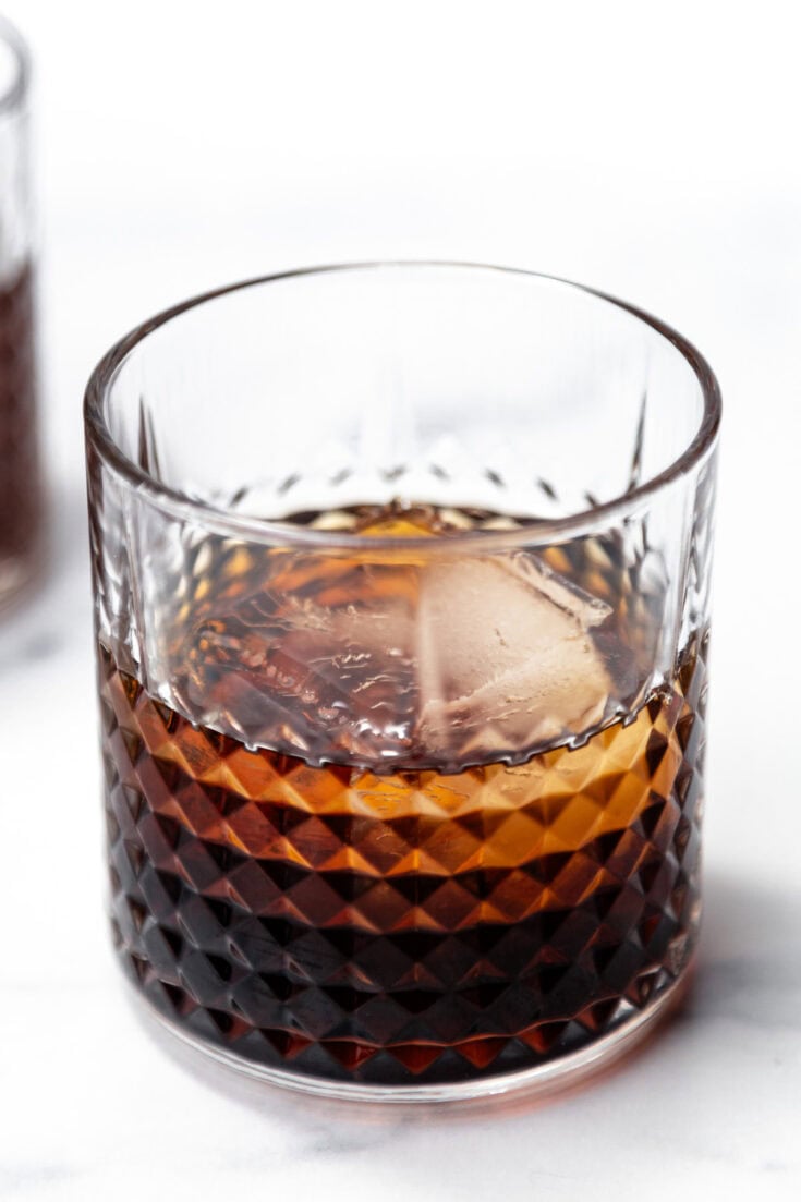 A glass of dark liquor with a large ice cube, set on a white surface.
