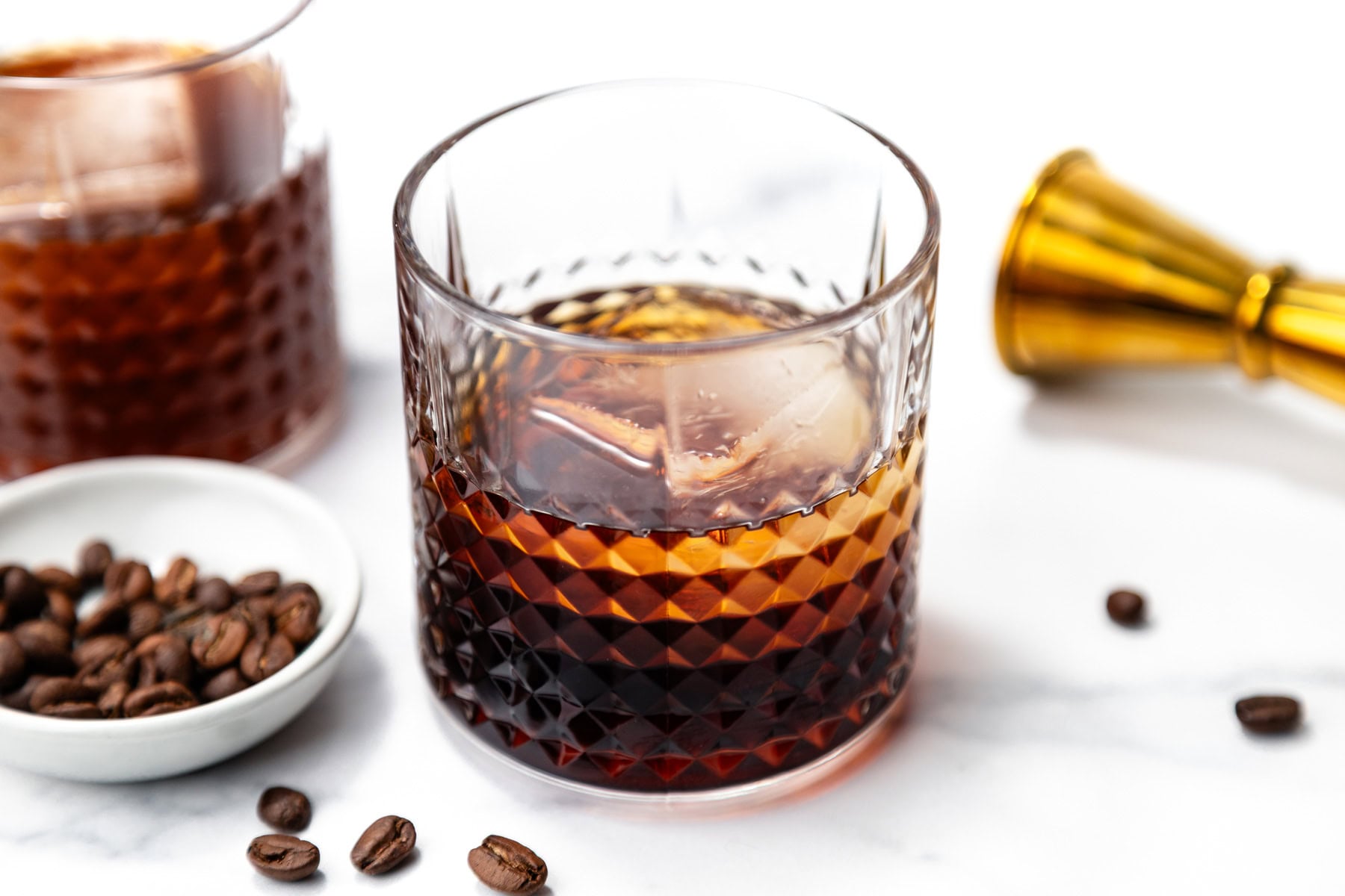 A glass of dark cocktail with ice sits on a white surface next to a dish of coffee beans, another glass, and a gold jigger.