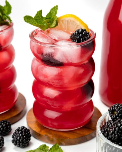 A glass of bright red iced drink garnished with a lemon slice, mint sprig, and blackberry, sits on a wooden coaster beside fresh blackberries and a matching bottle.
