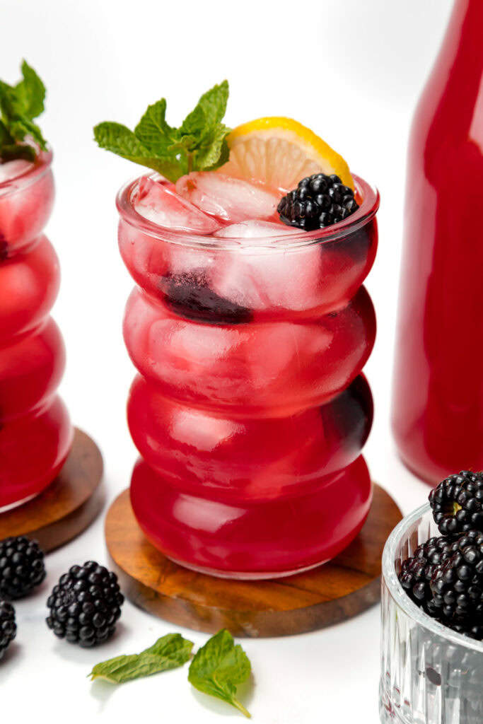 A glass of bright red iced drink garnished with a lemon slice, mint sprig, and blackberry, sits on a wooden coaster beside fresh blackberries and a matching bottle.