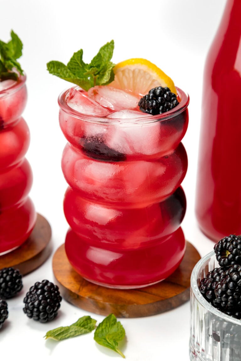 A glass of bright red iced drink garnished with a lemon slice, mint sprig, and blackberry, sits on a wooden coaster beside fresh blackberries and a matching bottle.