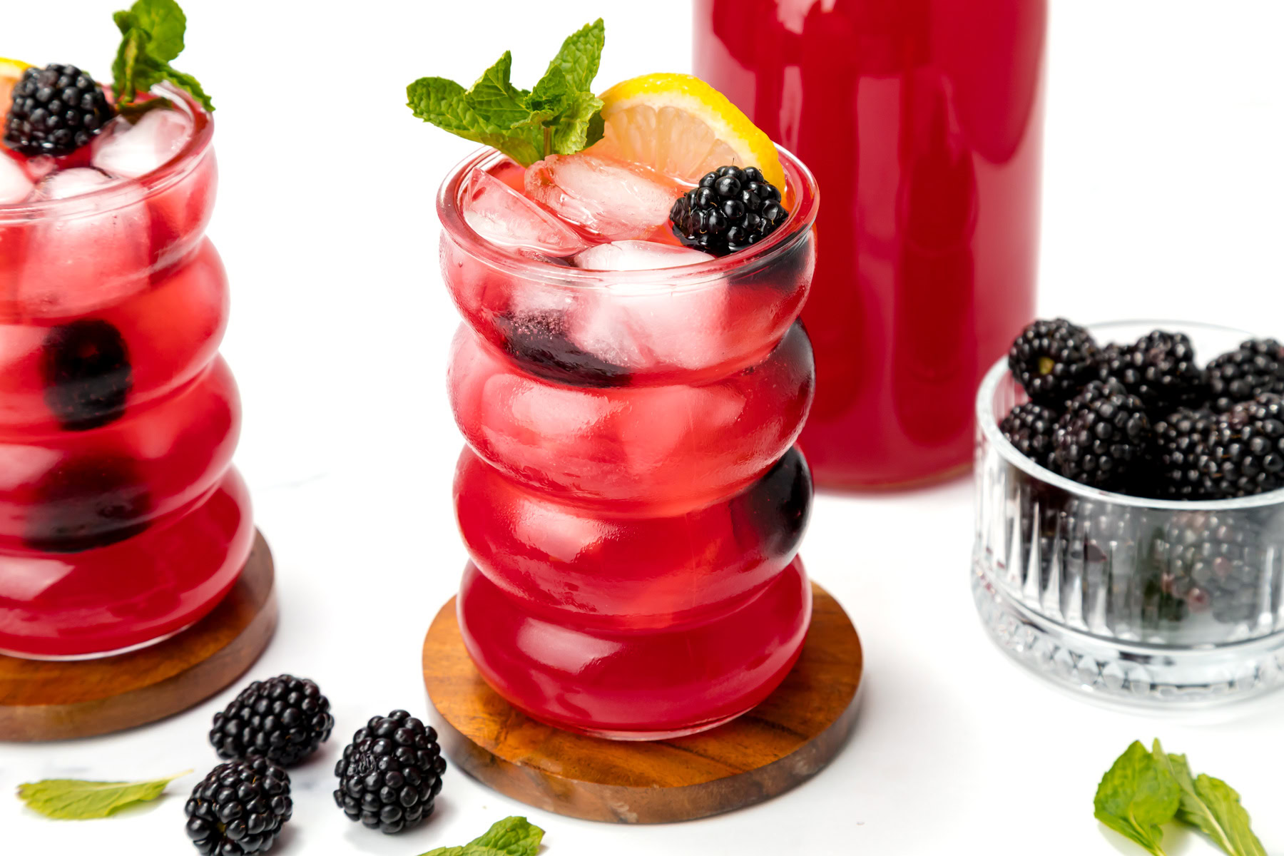 Two glasses of iced blackberry lemonade garnished with lemon, mint, and blackberries, with a bowl of blackberries and a bottle of the drink in the background.