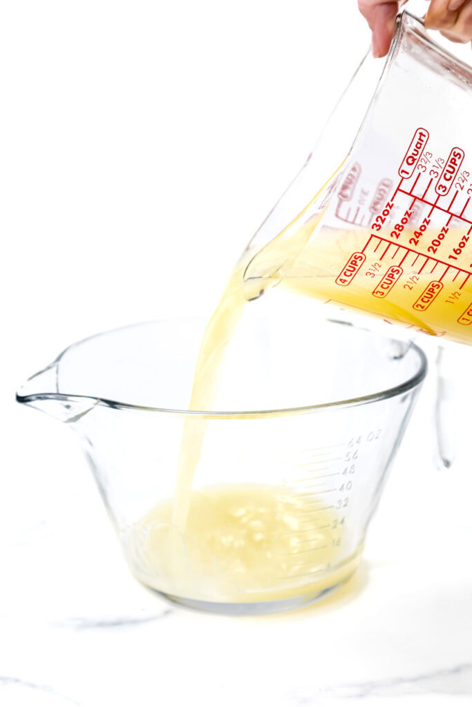 A hand pours a yellow liquid from a measuring cup into a larger clear glass measuring bowl on a white surface.