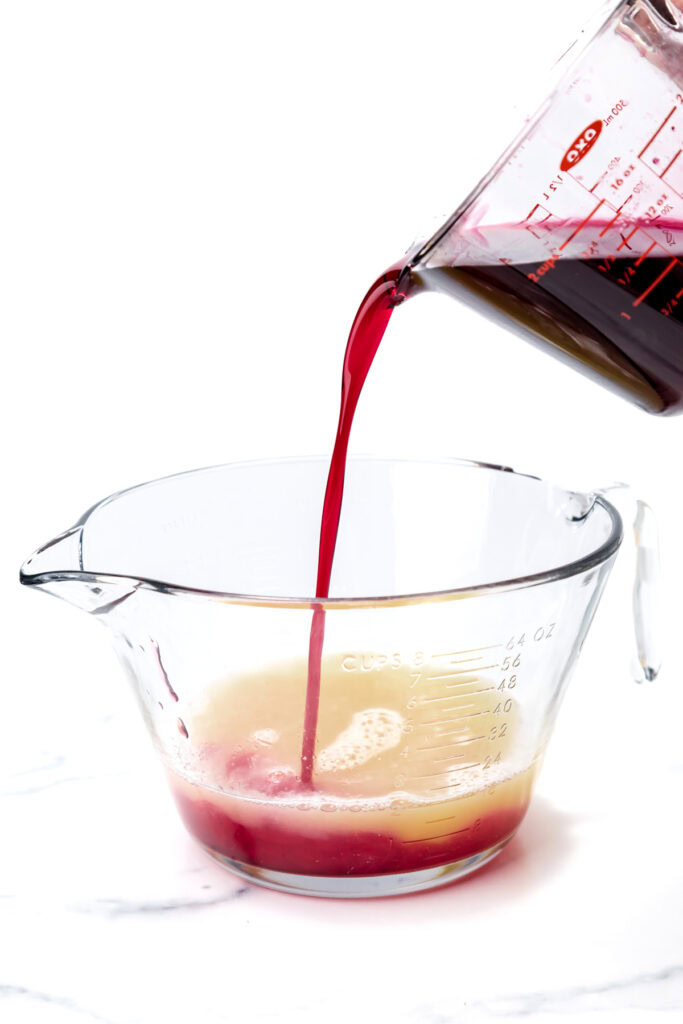 Red liquid is being poured from a measuring cup into another glass measuring cup containing a yellow liquid, against a white background.