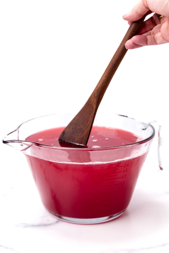 A hand stirs a red liquid in a large glass measuring cup with a wooden spatula.