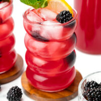 A red iced drink garnished with a lemon slice, mint sprig, and blackberry, served in a wavy glass on a wooden coaster.
