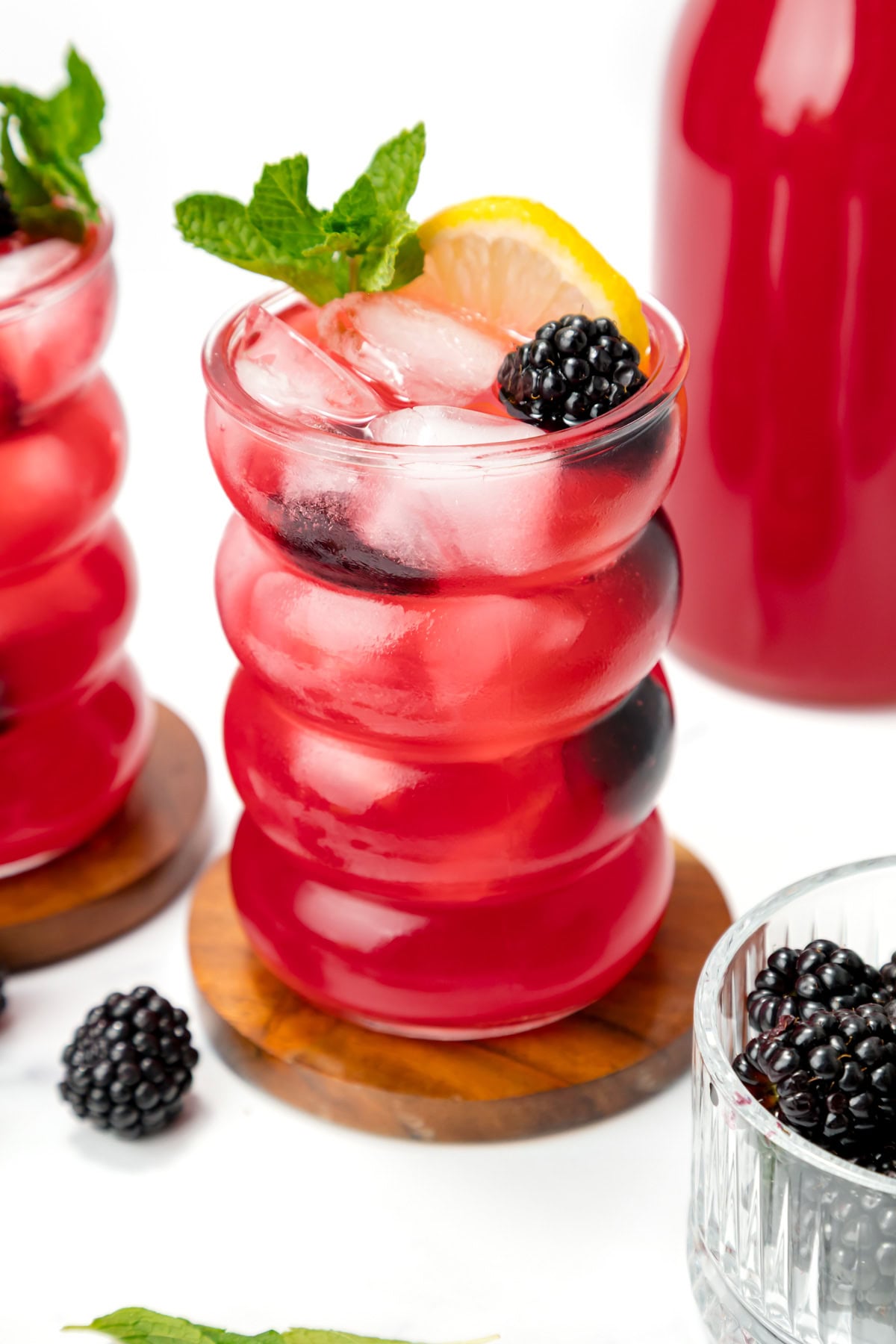 A red iced drink garnished with a lemon slice, mint sprig, and blackberry, served in a wavy glass on a wooden coaster.