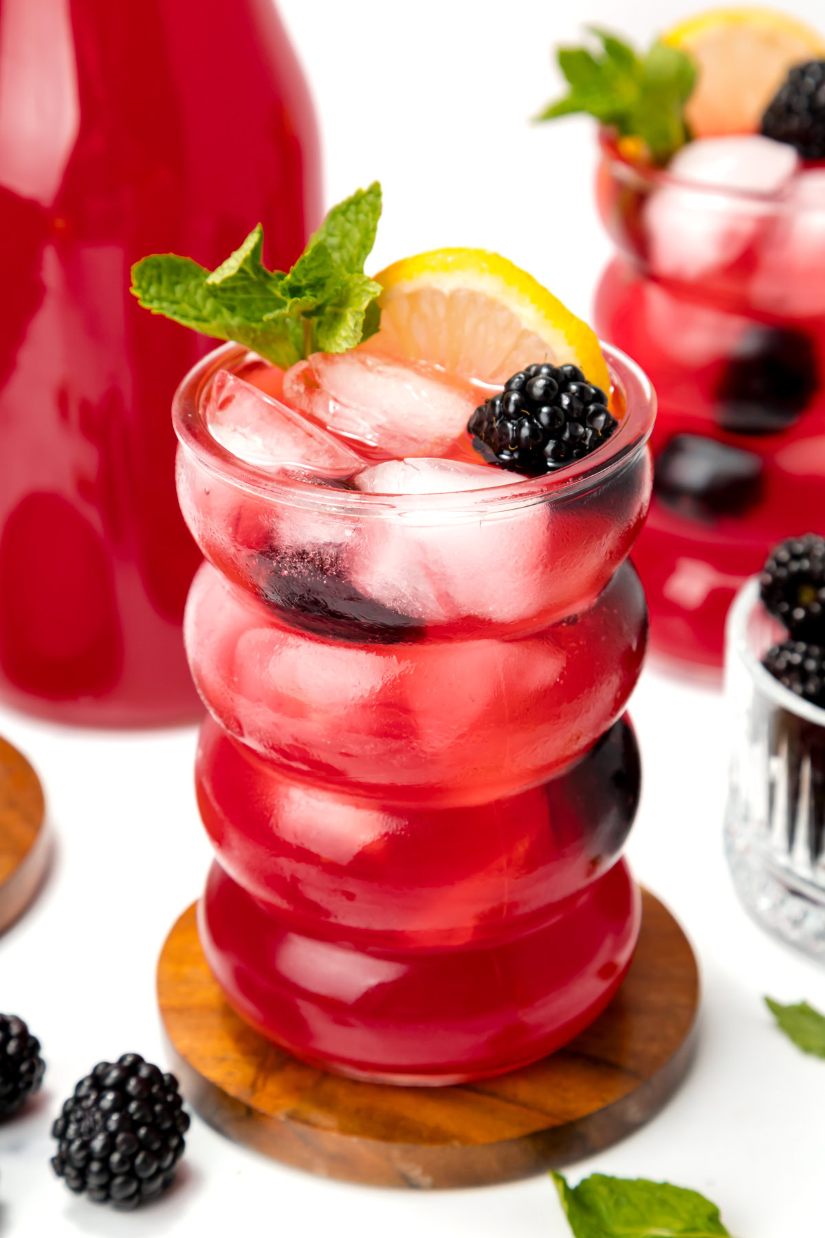 A glass of iced red berry drink garnished with a lemon slice, blackberry, and mint, with more berries and a drink pitcher in the background.