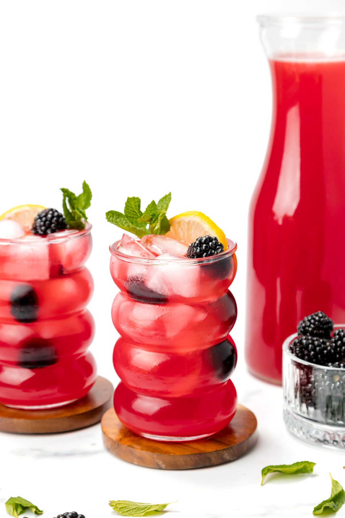 Two glasses of iced red berry lemonade garnished with lemon slices, blackberries, and mint, next to a carafe of the same drink and a small bowl of blackberries on a white background.