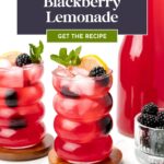 Two glasses of blackberry lemonade with ice, mint, lemon slices, and blackberries, next to a pitcher and a small bowl of blackberries on a white background.