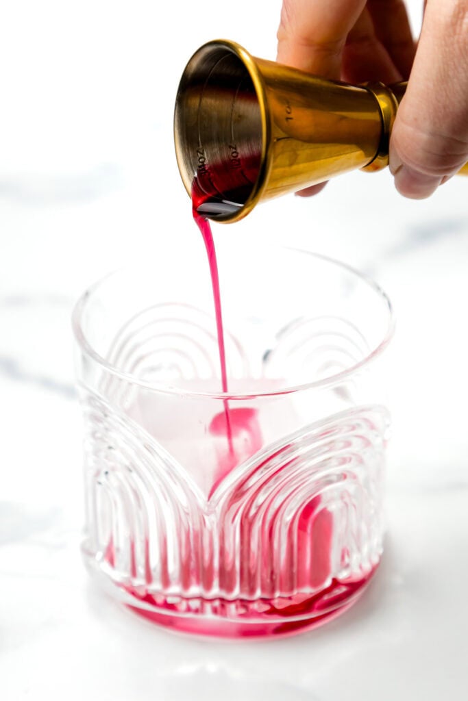 A hand pours a red liquid from a jigger into a patterned glass on a white surface.