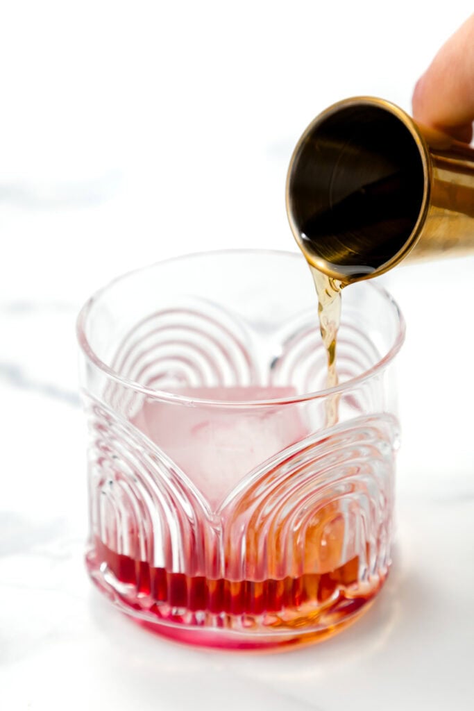 A hand pours liquid from a gold jigger into a glass with a pink-red beverage on a white surface.