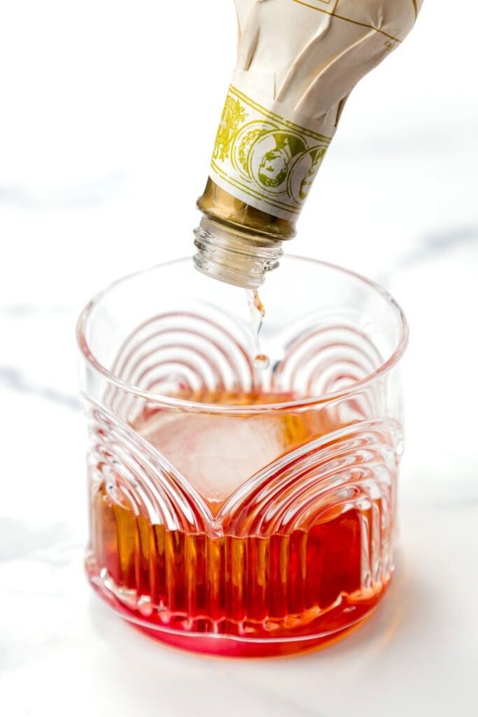 A bottle of bitters is being added to a glass of reddish-brown liquid on a white surface.
