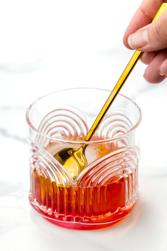 A hand stirs an amber-colored liquid in a patterned glass with a gold spoon on a white surface.