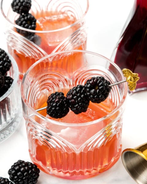 Two glasses of pink cocktail with ice, garnished with skewered blackberries, sit on a white surface alongside fresh blackberries and a cocktail shaker.