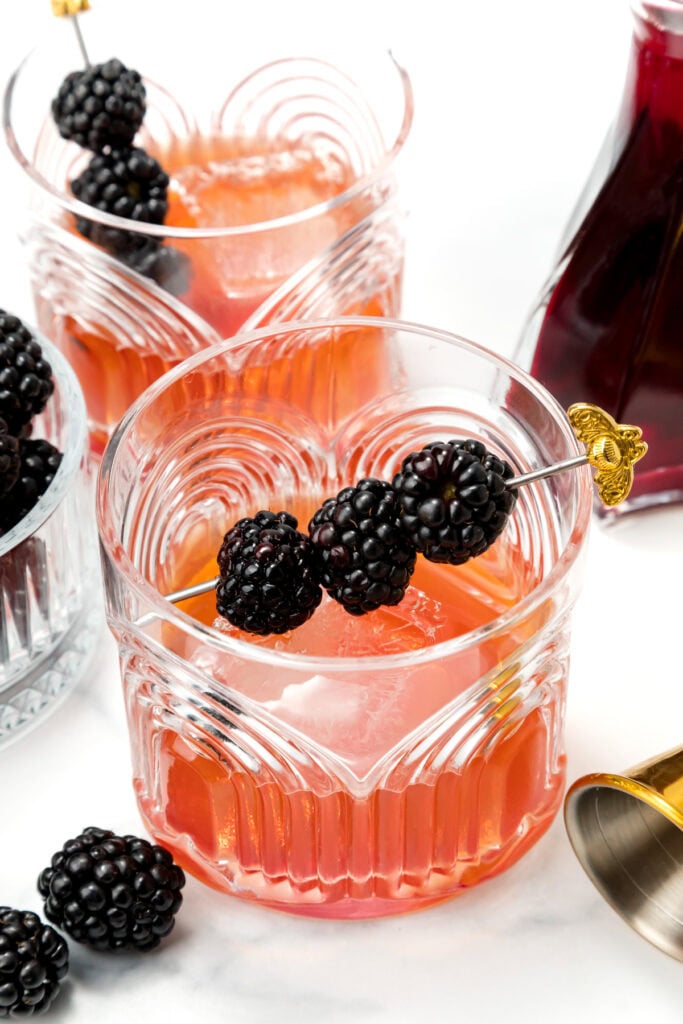 Two glasses of pink cocktail with ice, garnished with skewered blackberries, sit on a white surface alongside fresh blackberries and a cocktail shaker.