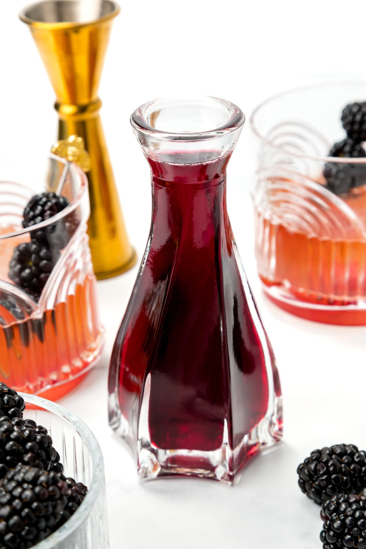 A glass carafe filled with dark red liquid is surrounded by blackberries, two cocktail glasses, and a gold jigger on a white surface.