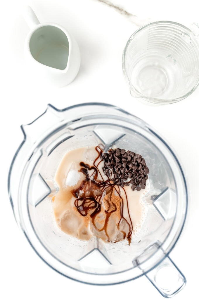 Top-down view of a blender containing ice, chocolate syrup, chocolate chips, and milk, with a small pitcher and an empty measuring cup nearby on a white surface.