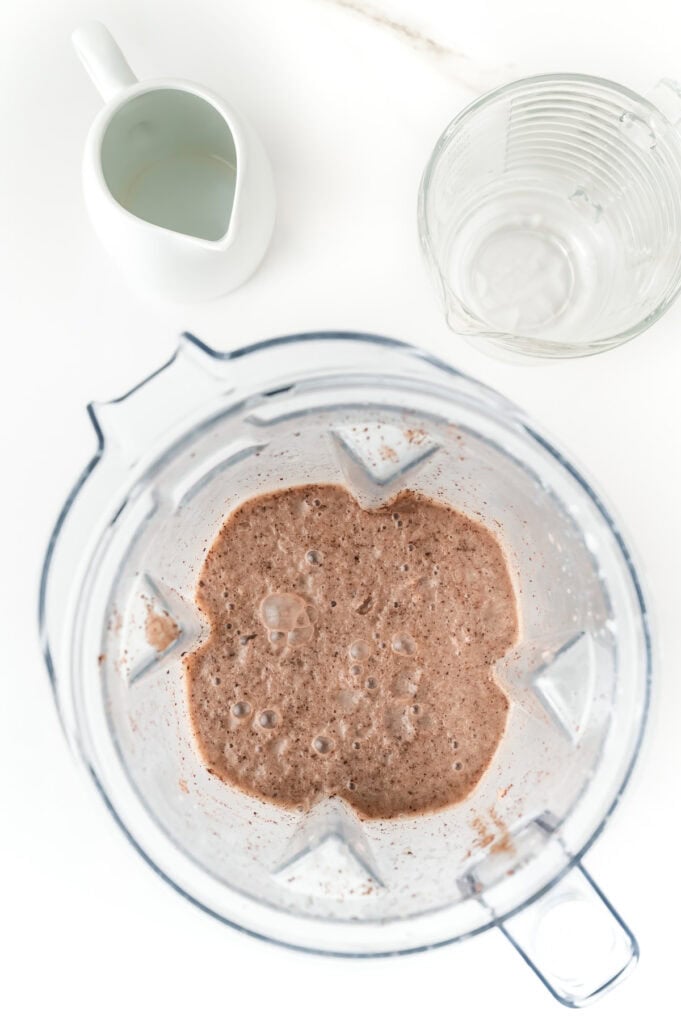 Top view of a blender containing a frothy chocolate mixture, next to an empty glass measuring cup and a small white pitcher on a white surface.