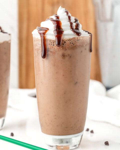 A glass of chocolate milkshake topped with whipped cream and chocolate syrup, with chocolate chips and a green straw on the table.