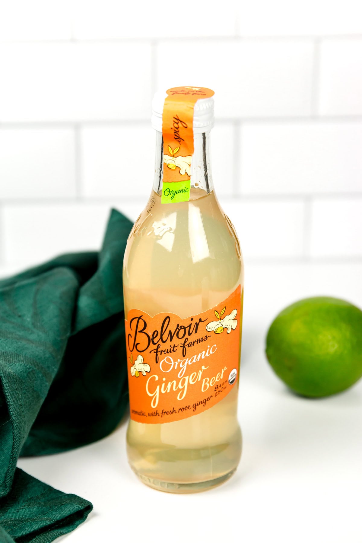 A bottle of Belvoir Fruit Farms Organic Ginger Beer stands on a white surface next to a green cloth and a whole lime, with a white tiled background.