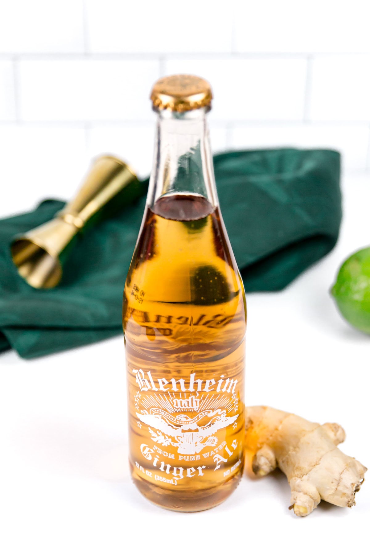 A bottle of Blenheim Ginger Ale is placed on a white surface with a piece of ginger, a green lime, a gold jigger, and a dark green cloth in the background.