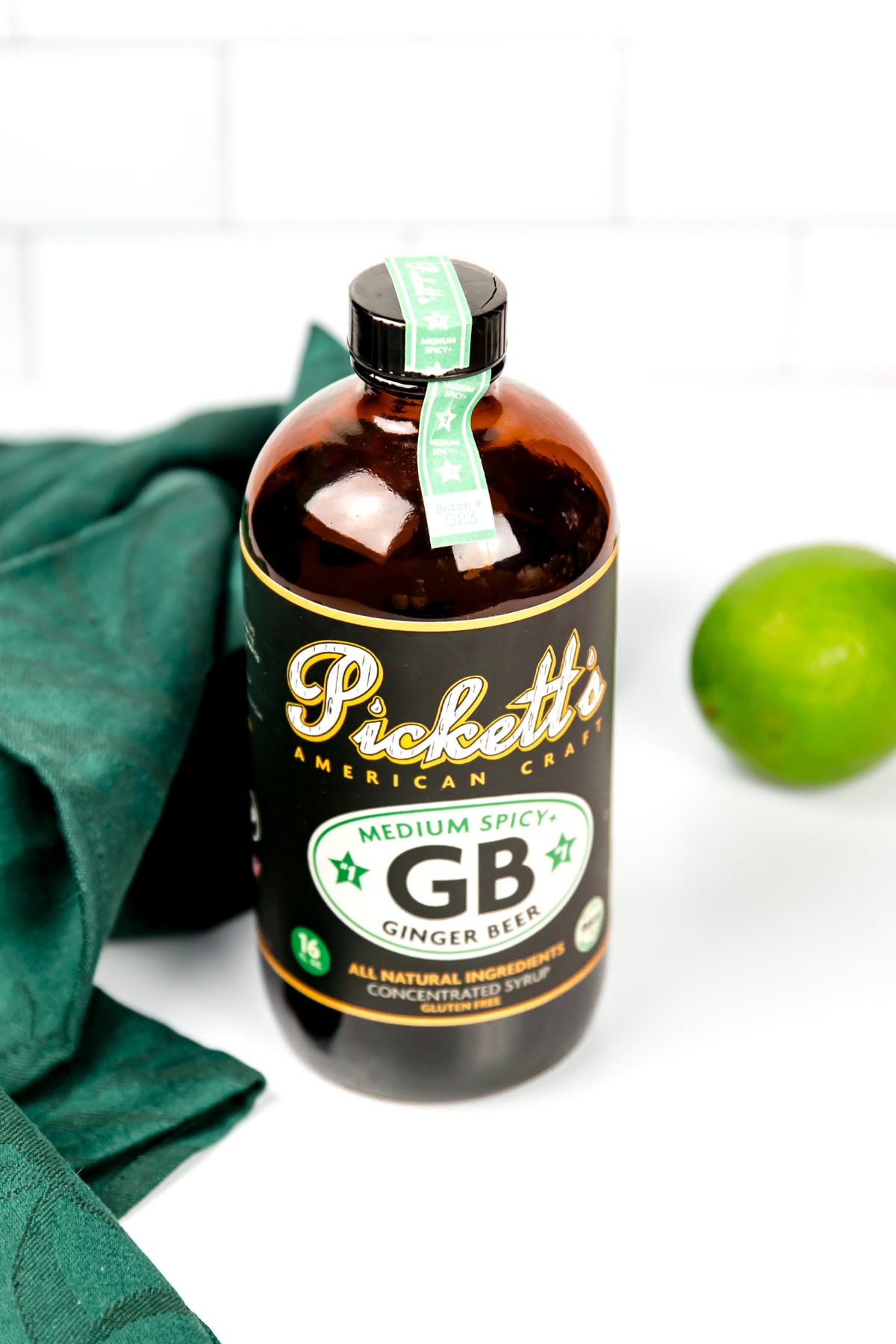 A bottle of Pickett’s Medium Spicy Ginger Beer concentrate sits on a white surface next to a green cloth and a lime.