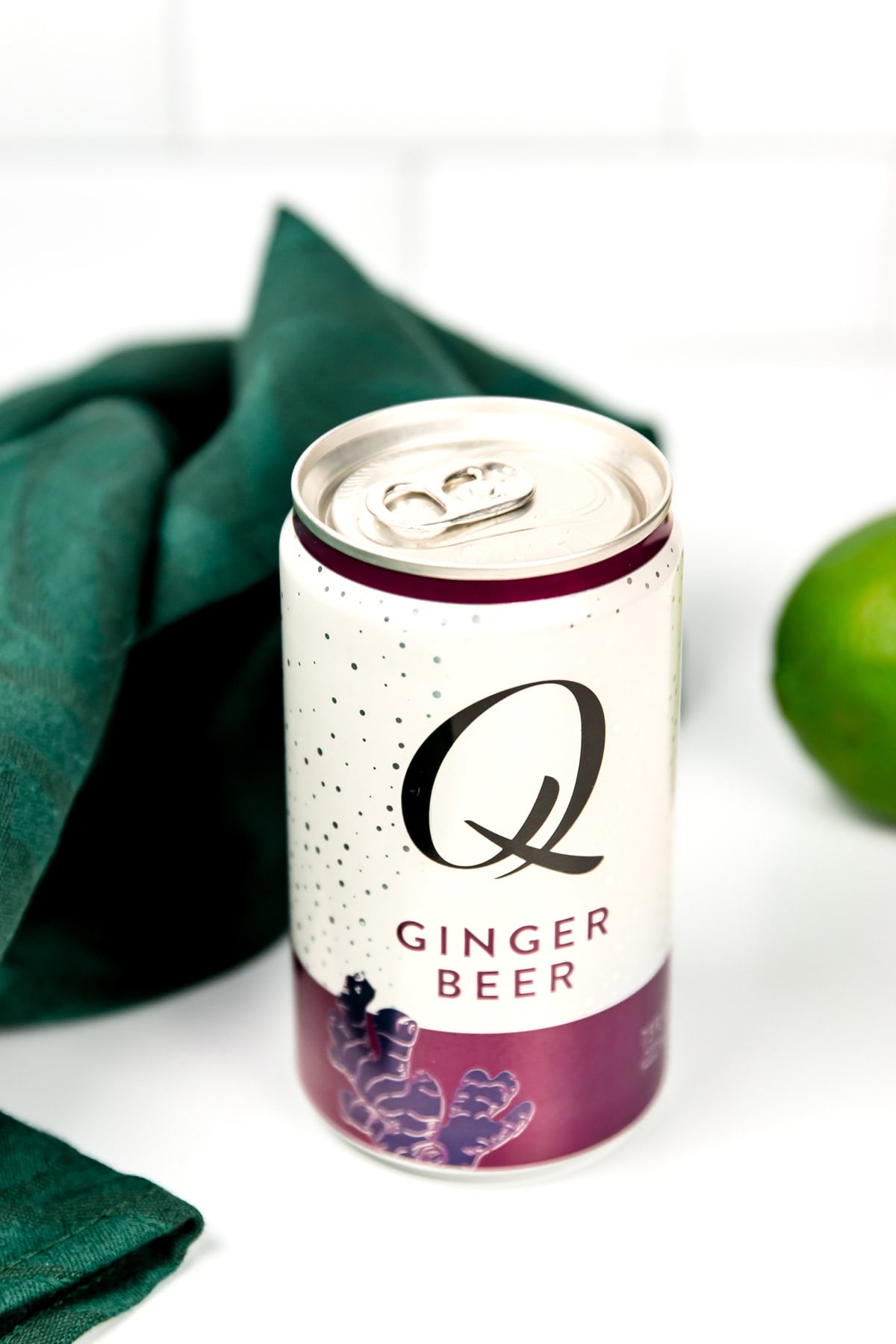 A can of Q Ginger Beer sits on a white surface next to a green cloth and a lime, with the tab opened.