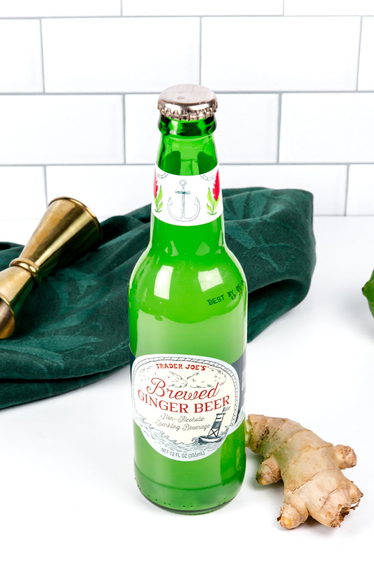 A glass bottle of Trader Joe’s Brewed Ginger Beer stands on a white surface with a piece of ginger, a green cloth, and a metal jigger nearby.