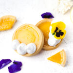 Two lemon tarts, one topped with whipped cream and the other with whipped cream and a yellow pansy, are displayed on a white surface with tart slices and purple flower petals.