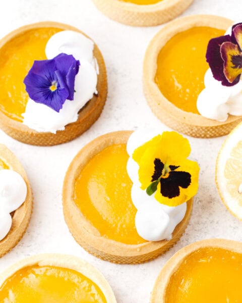Small lemon tarts with bright yellow filling, some topped with whipped cream and edible flowers, arranged on a white surface.