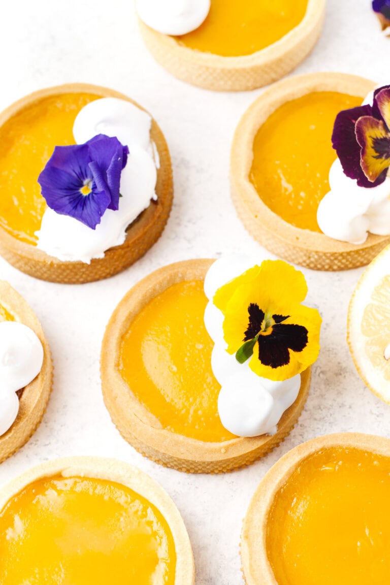 Small lemon tarts with bright yellow filling, some topped with whipped cream and edible flowers, arranged on a white surface.