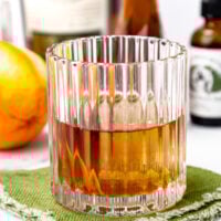 A glass of amber-colored liquor sits on a green textured napkin, with an orange and various bottles blurred in the background.