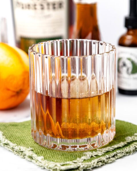 A glass of amber-colored liquor sits on a green textured napkin, with an orange and various bottles blurred in the background.