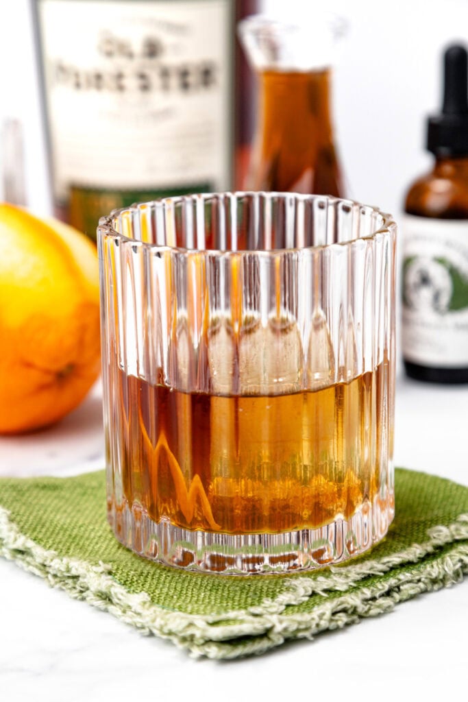 A glass of amber-colored liquor sits on a green textured napkin, with an orange and various bottles blurred in the background.