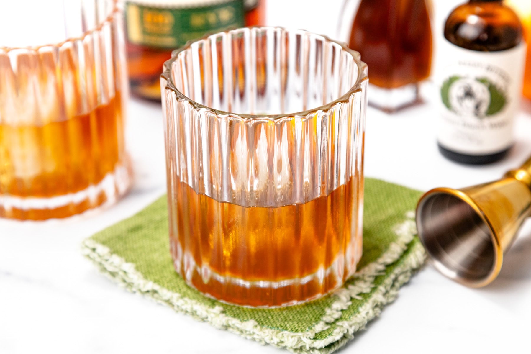 A glass of amber-colored cocktail sits on a green napkin, with a metal jigger, bitters bottle, and other glasses in the background.