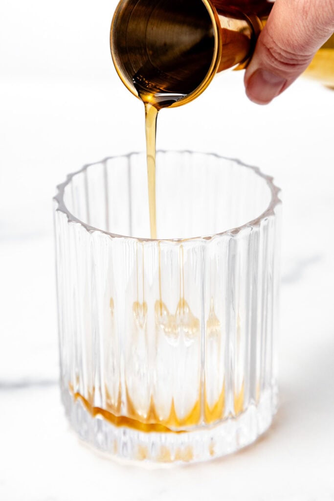 A hand pours a golden liquid from a metal jigger into a clear, textured glass on a white surface.