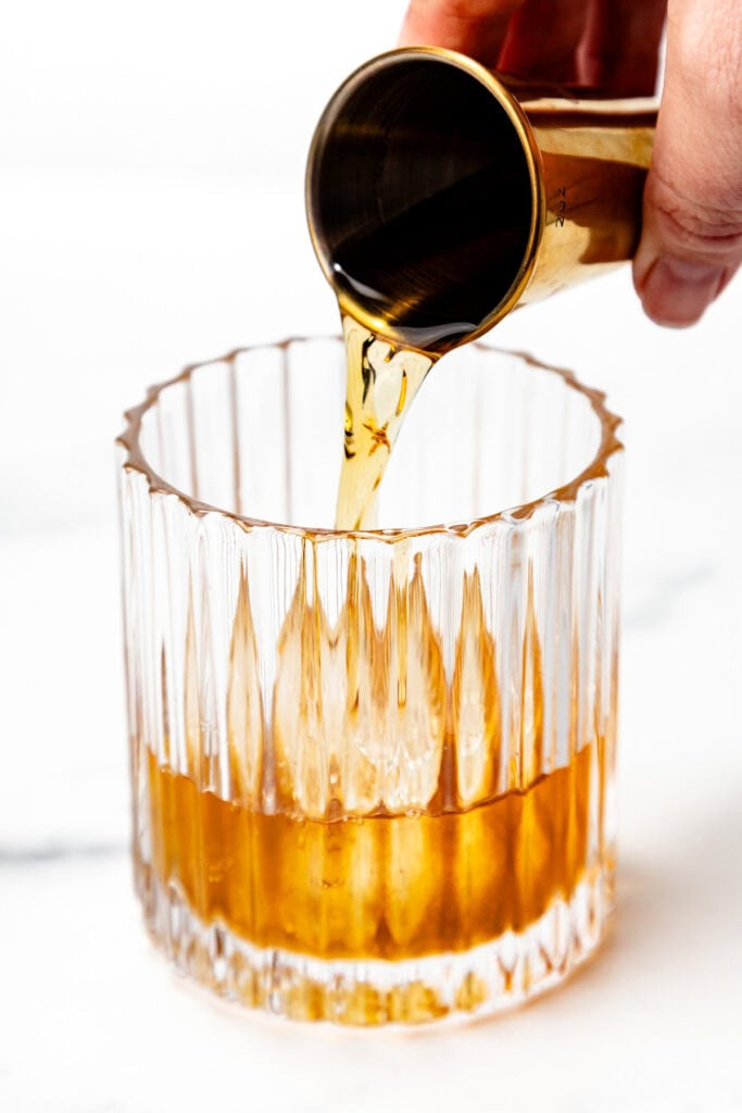 A hand pours amber liquid from a jigger into a faceted glass tumbler against a white background.