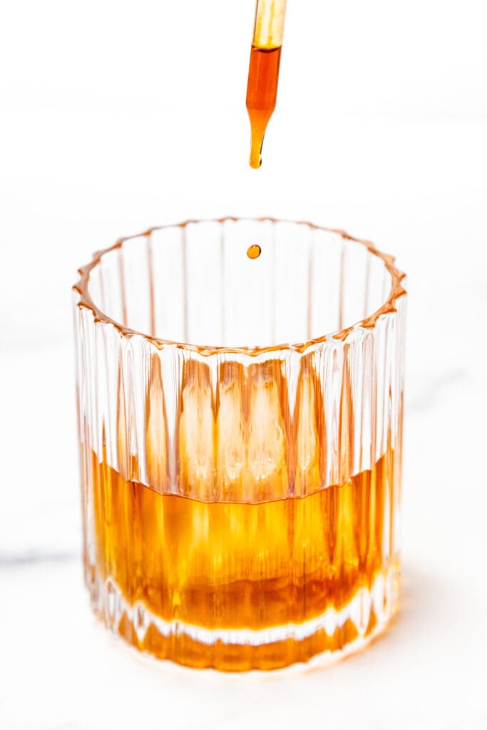 A dropper releases a droplet of liquid into a glass filled with an amber-colored beverage.