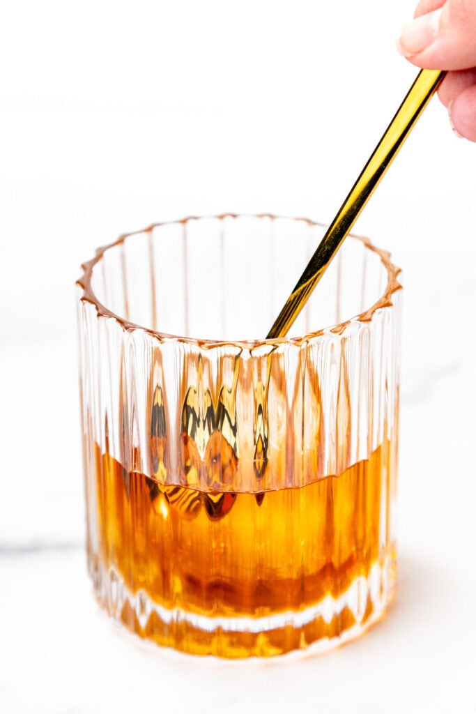 A hand stirs an amber-colored drink in a faceted glass with a gold spoon on a white background.