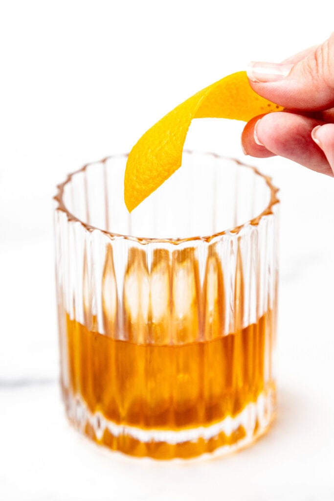A hand holds an orange peel above a glass of amber-colored liquid, possibly a cocktail, on a white background.