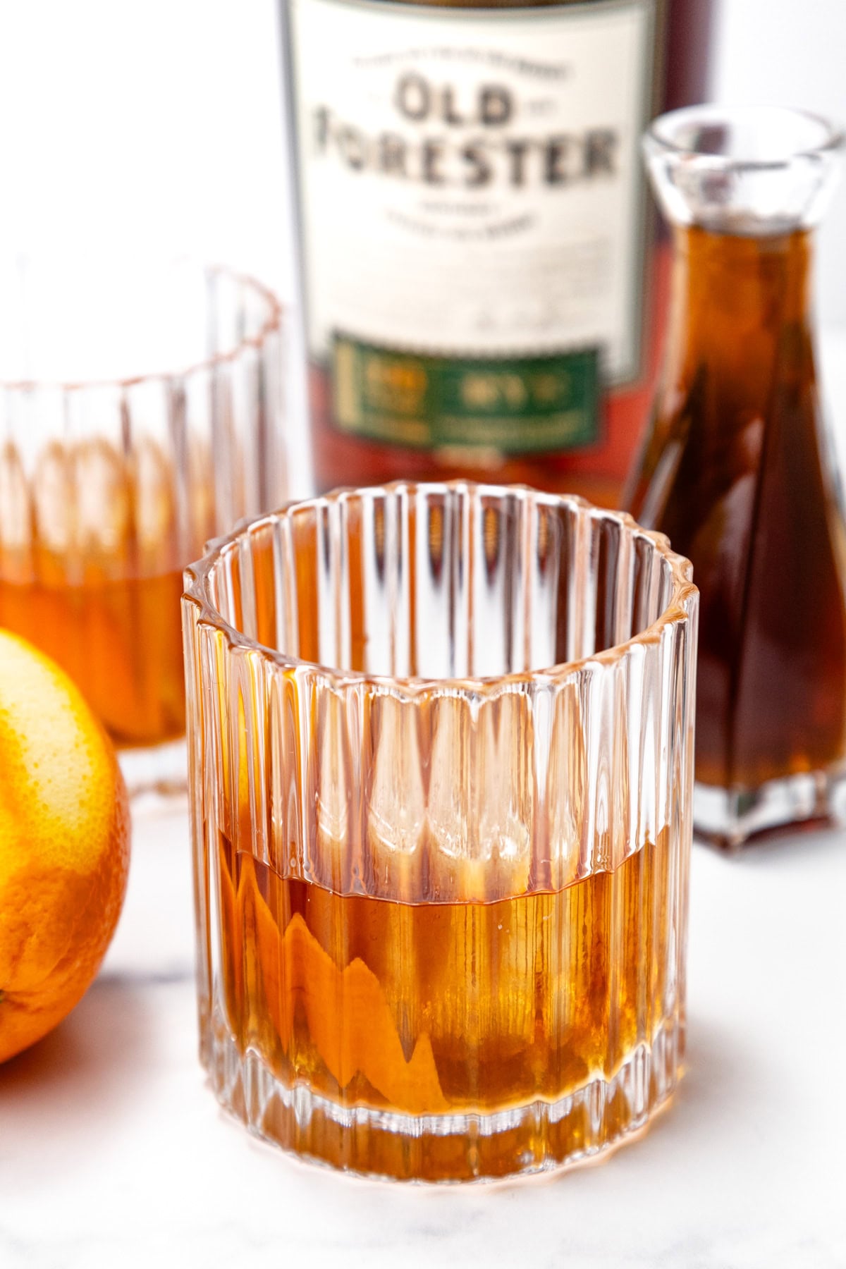 A glass of whiskey on ice sits on a table with an orange, a small carafe of brown liquid, and a bottle labeled "Old Forester" in the background.