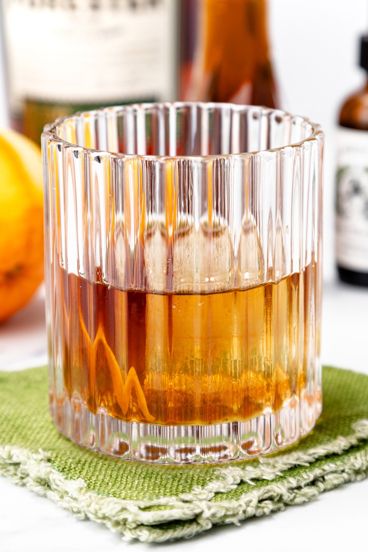 A glass of amber-colored cocktail with a large ice cube, placed on a green napkin, with an orange and bottles blurred in the background.