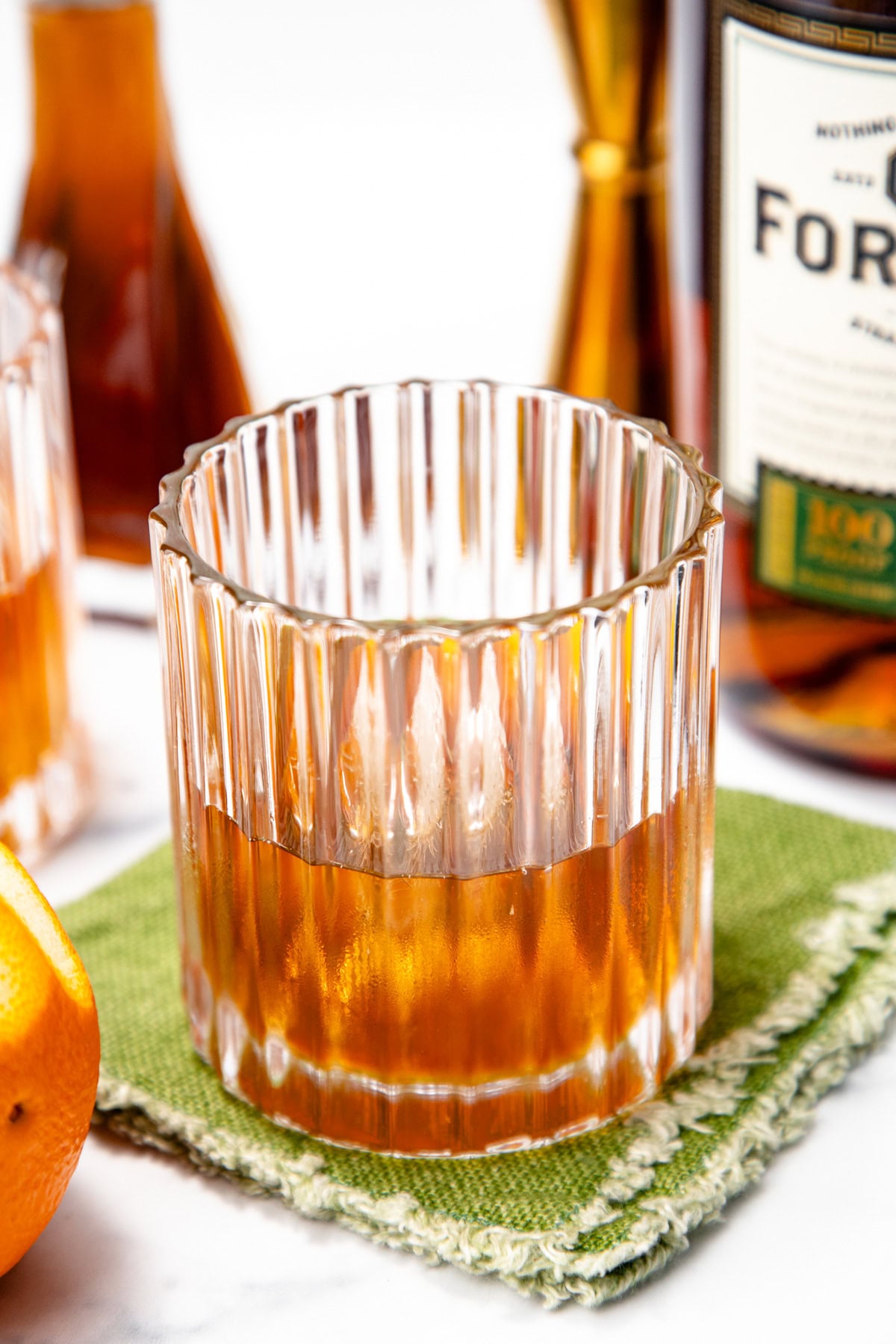 A glass of whiskey sits on a green textured napkin, with an orange and a whiskey bottle in the background.