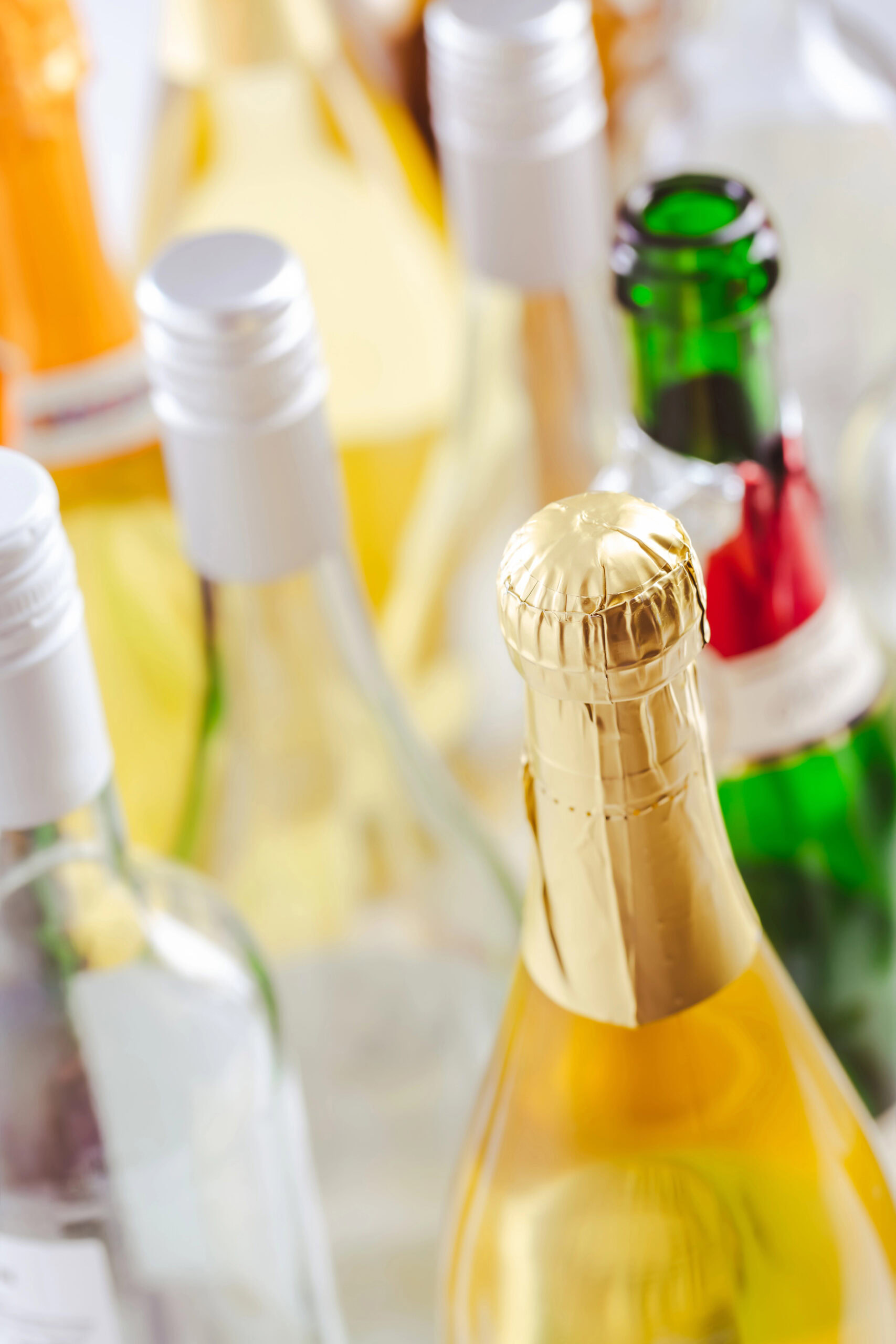 Assorted bottles of alcoholic beverages, including wine and champagne, with various colored caps and foil tops, are grouped closely together.