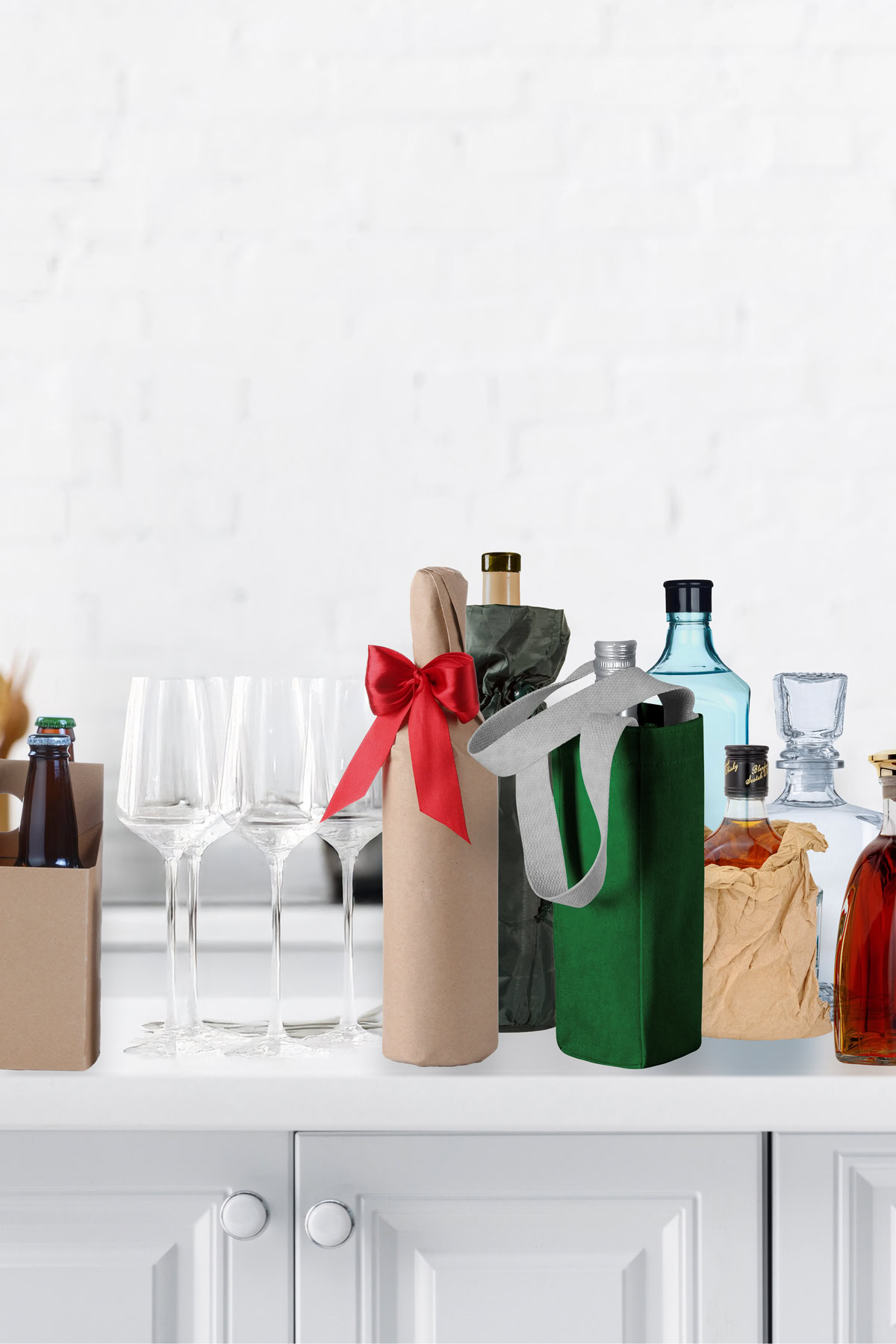 A variety of bottled drinks and gift-wrapped bottles are arranged on a white countertop, with four empty wine glasses beside them.